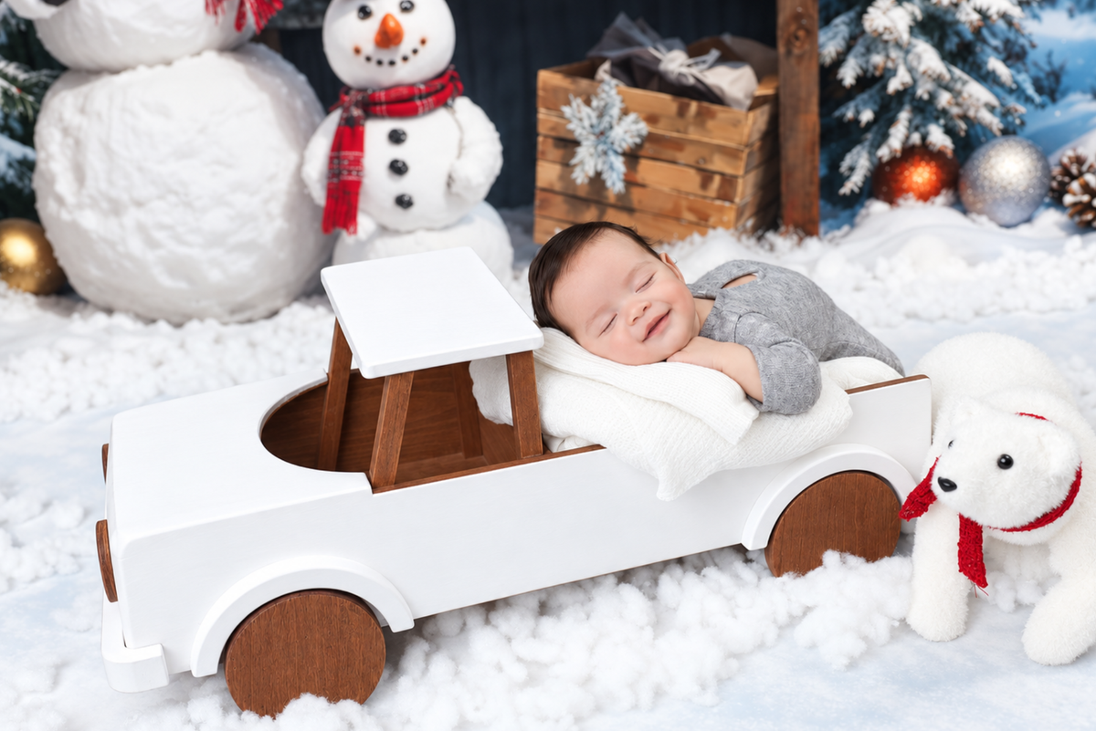 A sleeping baby lies in the bed of a white wooden pickup truck decorated with brown wood panels, posed in a snowy Christmas-themed studio scene with a snowman, tree, ornaments, and a plush polar bear. Great for newborn photography