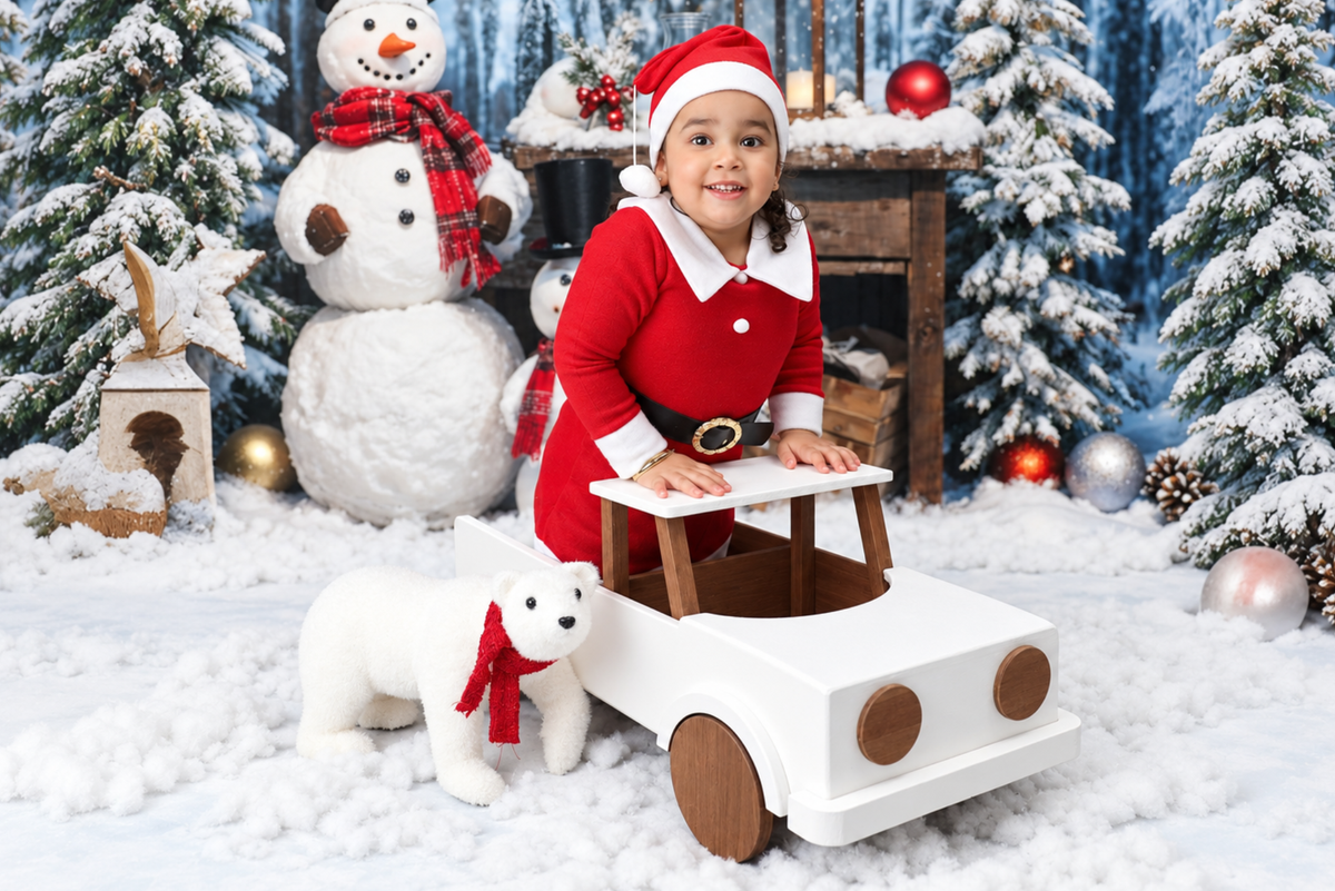 A young child dressed in a red Santa outfit and Santa hat sits in a white wooden pickup truck prop, surrounded by a snowy winter holiday scene with a snowman, decorated trees, ornaments, and a plush polar bear.