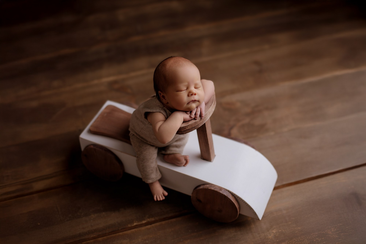 Newborn posed on Rustic Ride-On, a wooden car-shaped newborn photography prop, with warm wood floor.