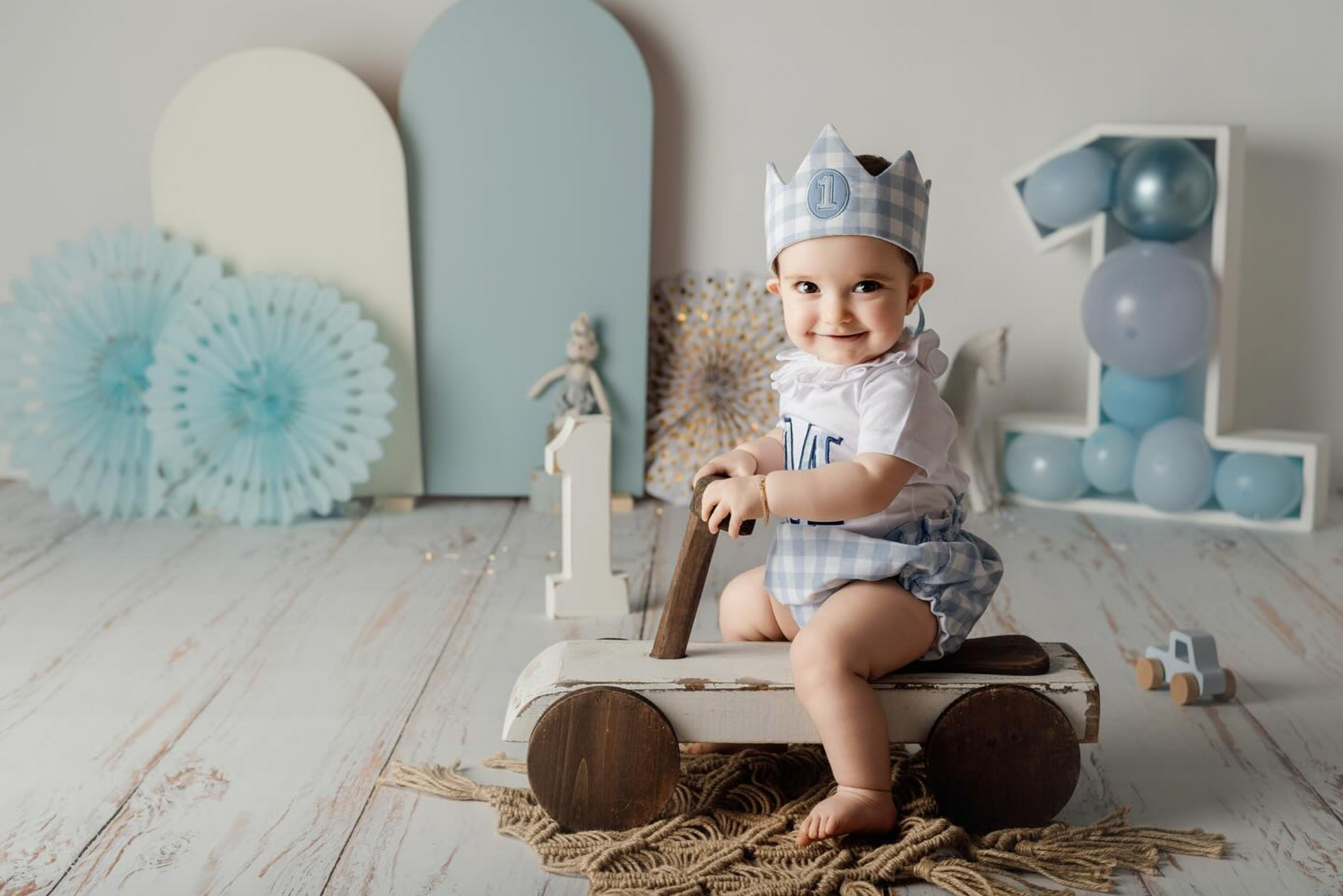 Baby wearing a gingham crown and romper smiles while sitting on a wooden ride-on car, styled with blue paper fans, balloon decor, and a soft rustic mat; perfect newborn photography prop for milestone portraits