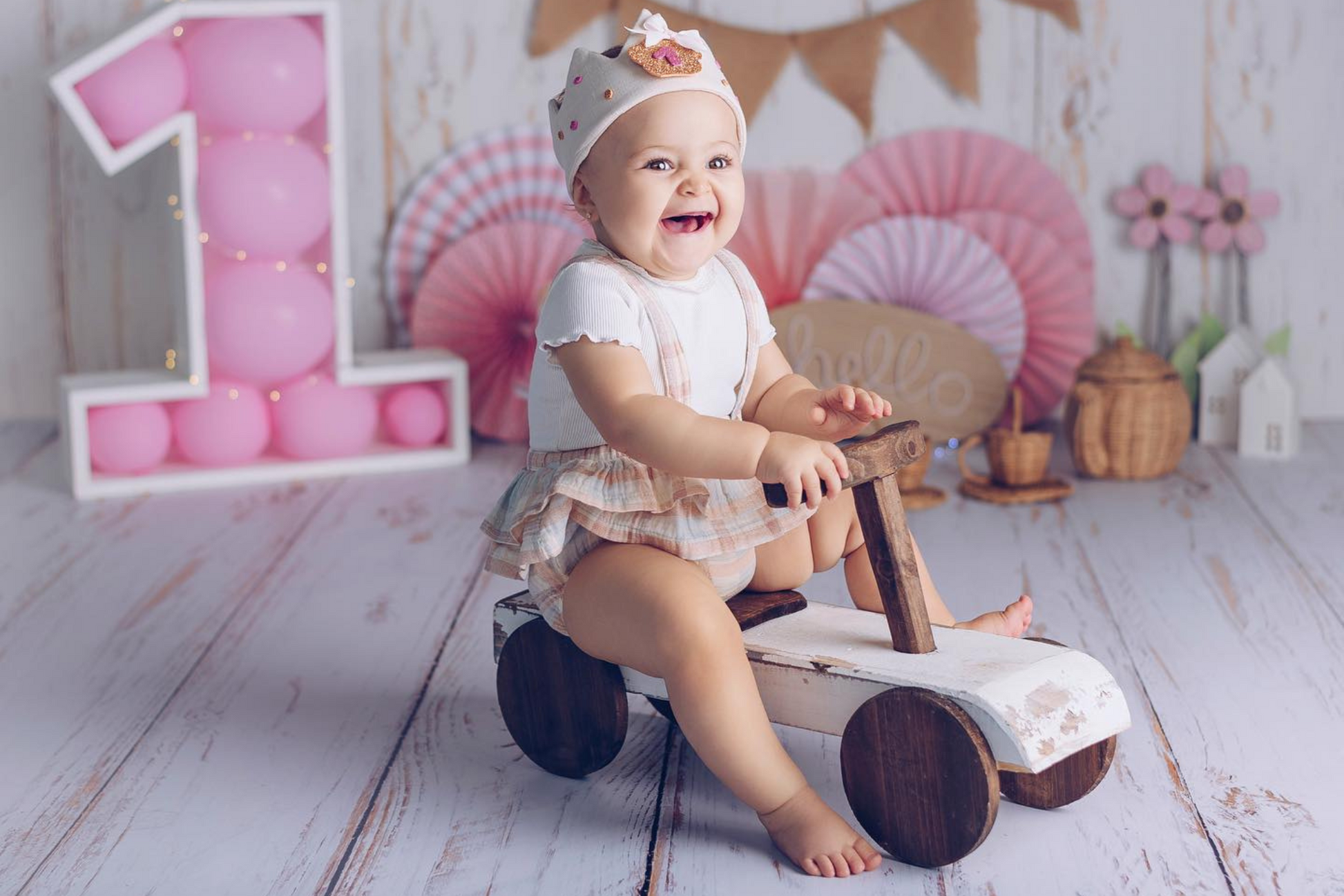 Smiling baby in a white outfit and crown sits on a rustic wooden ride-on car, surrounded by pink decor, including a balloon-filled number one; ideal newborn photography prop for milestone sessions.