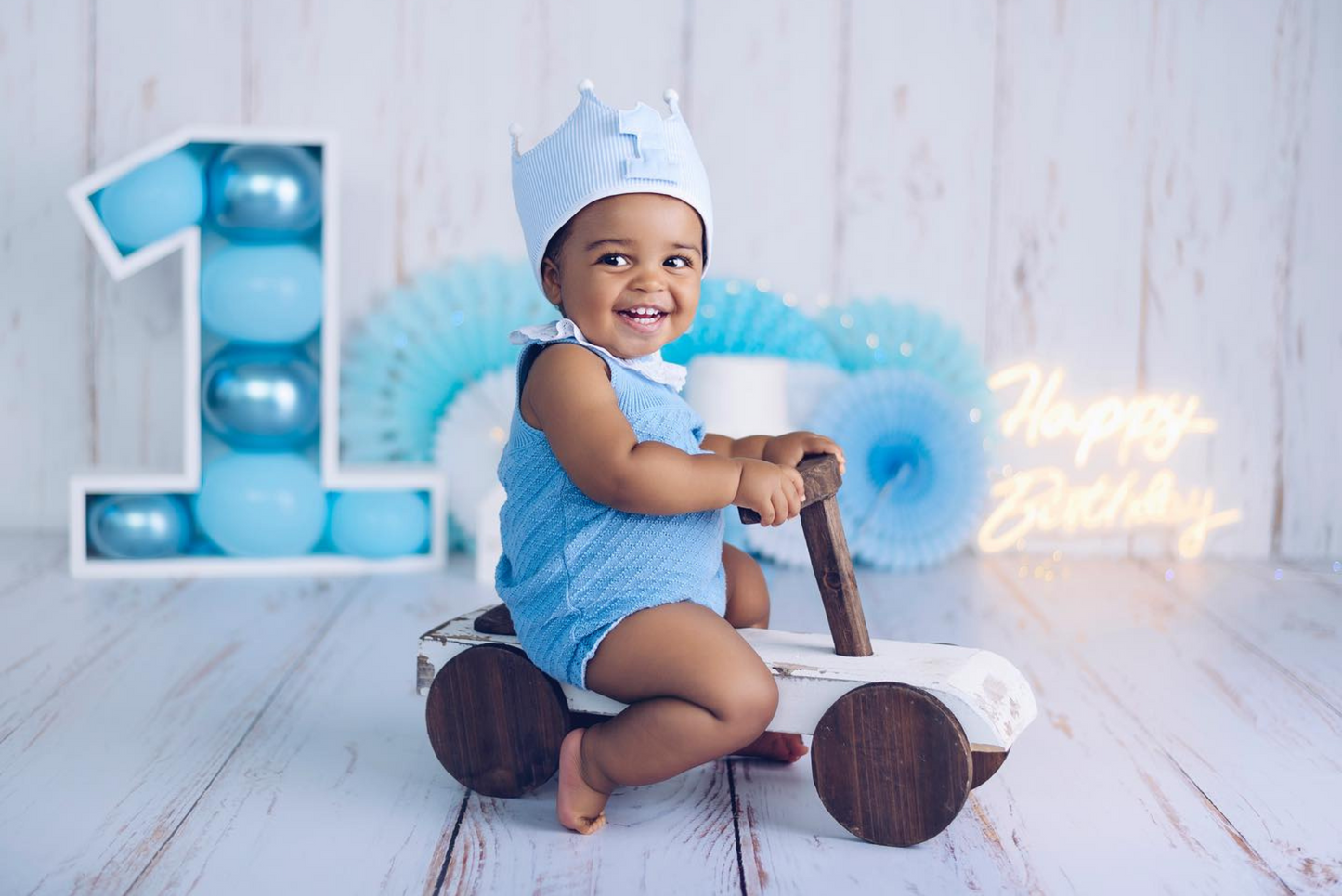 Cheerful baby in a blue romper and crown rides a vintage wooden prop car, set against a blue-themed first birthday backdrop with a balloon-filled number one and glowing "Happy Birthday" sign