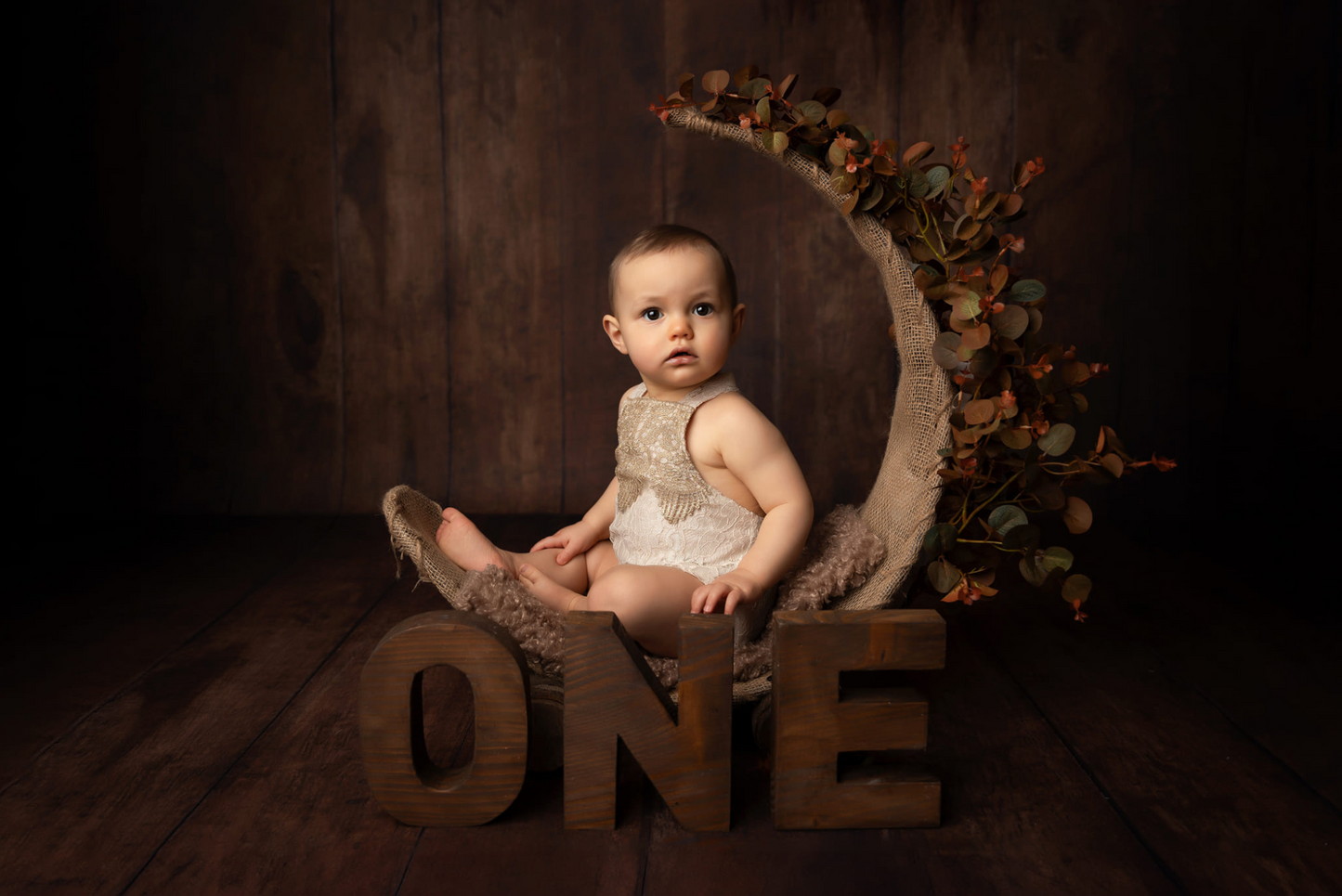 A baby sits in a rustic moon-shaped swing adorned with autumn leaves, with large wooden letters spelling "ONE" in front. The scene uses a newborn photography prop to create a warm, earthy atmosphere.