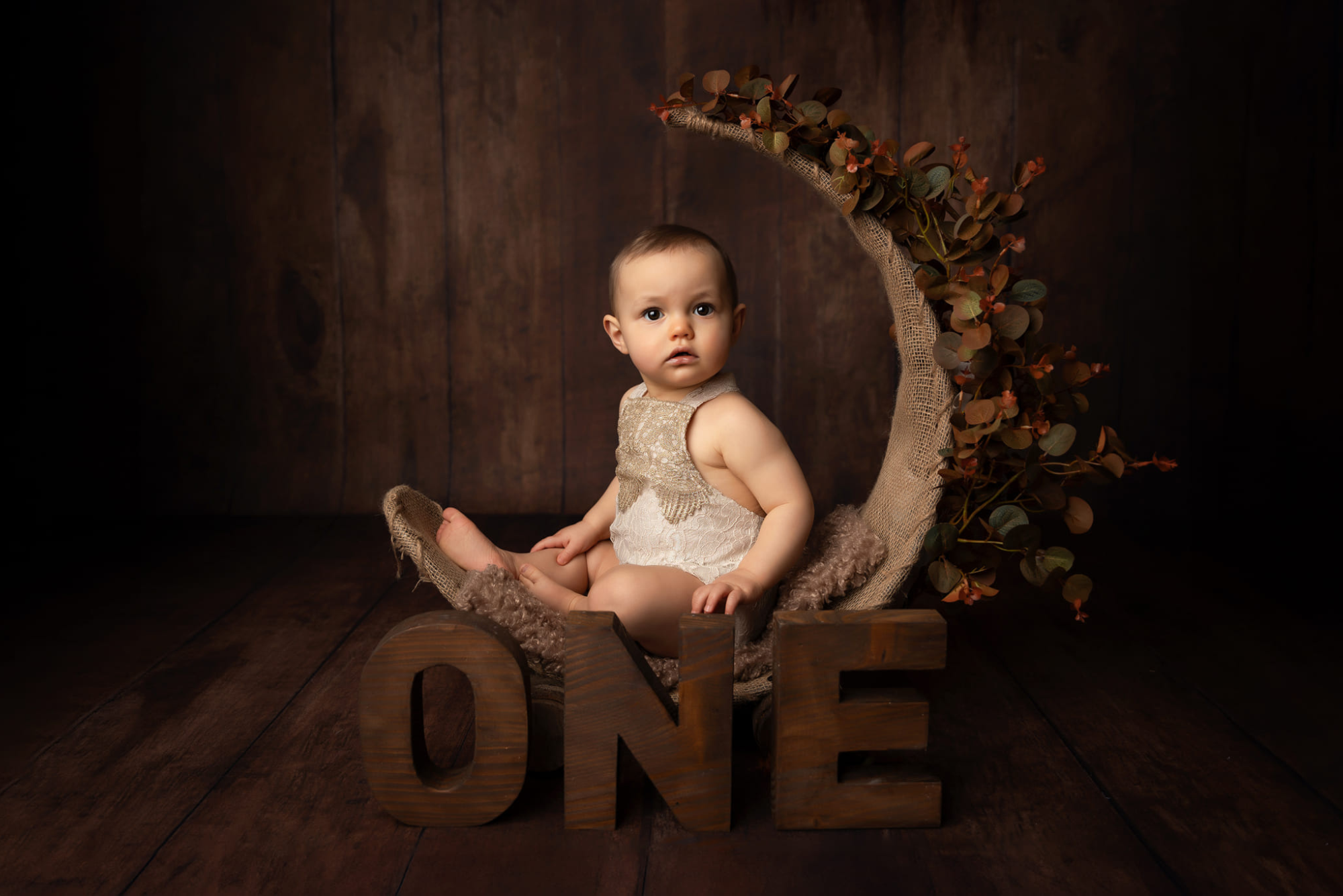 A baby sits in a rustic moon-shaped swing adorned with autumn leaves, with large wooden letters spelling "ONE" in front. The scene uses a newborn photography prop to create a warm, earthy atmosphere.