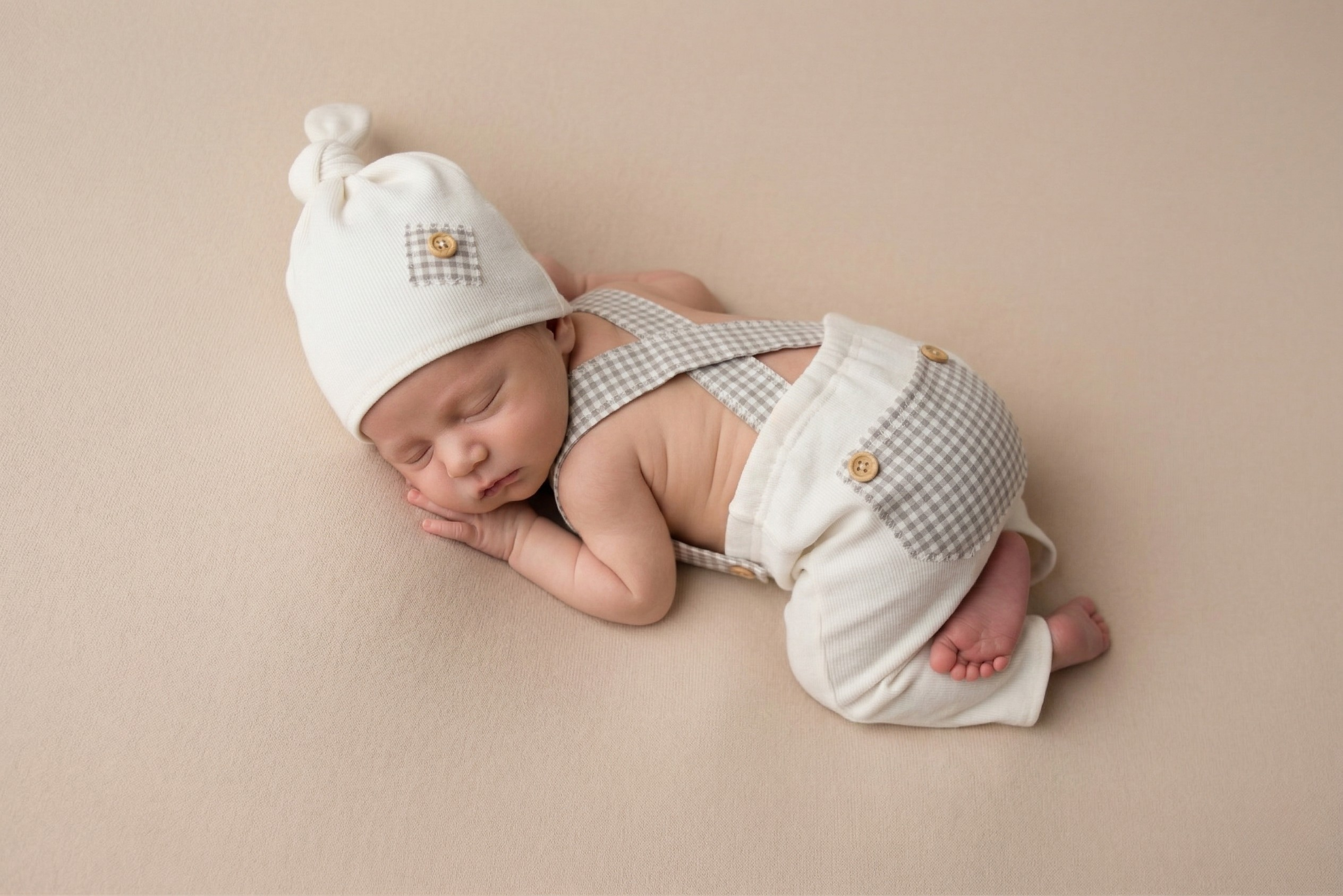 Newborn baby in white hat and checkered outfit lying on a beige surface