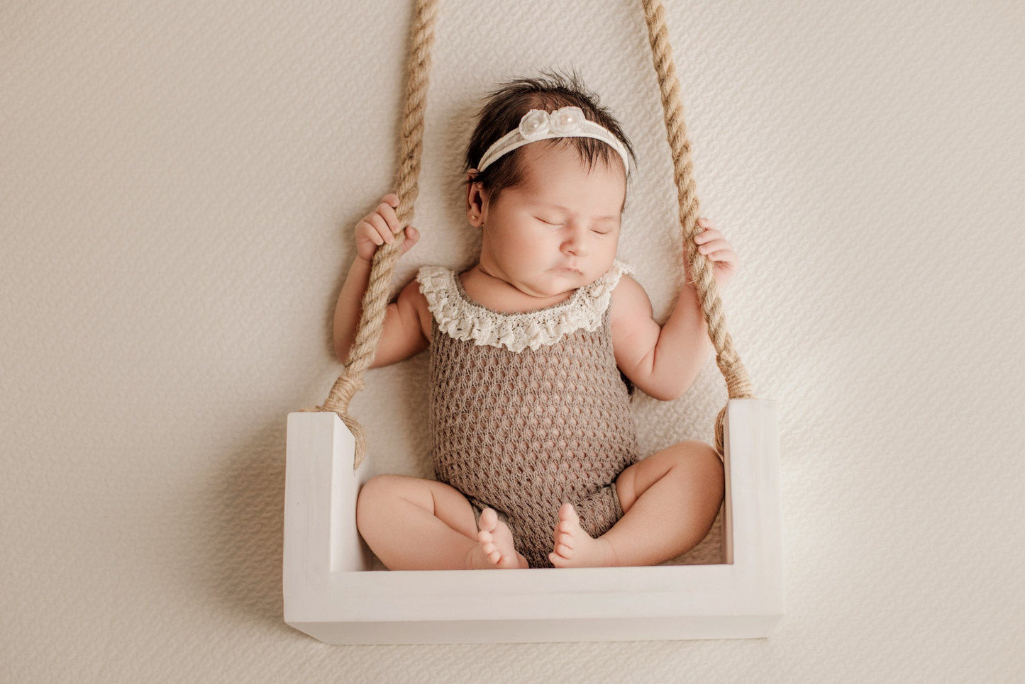 Newborn asleep on a white rustic swing, wearing a knit prop outfit. 