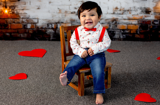 Adorable Valentine’s-themed setup featuring a barefoot toddler sitting on a Small Wooden Harlow Chair, dressed in a heart-patterned shirt, red suspenders, and a bow tie. Red heart decorations are scattered on the floor, with a rustic brick and whitewashed wall backdrop. Perfect newborn photography prop for milestone sessions!