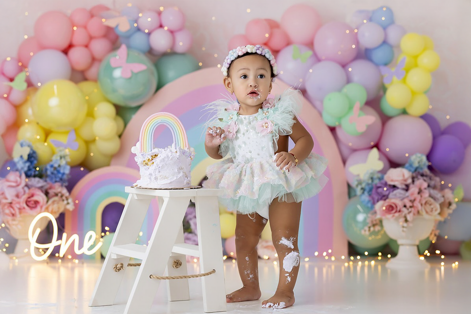 Adorable baby girl in a pastel tutu dress stands beside a white wooden step ladder mini, featuring a rainbow-themed smash cake. The whimsical newborn photography prop is set against a dreamy pastel balloon backdrop with butterflies and fairy lights, creating a magical first birthday scene.