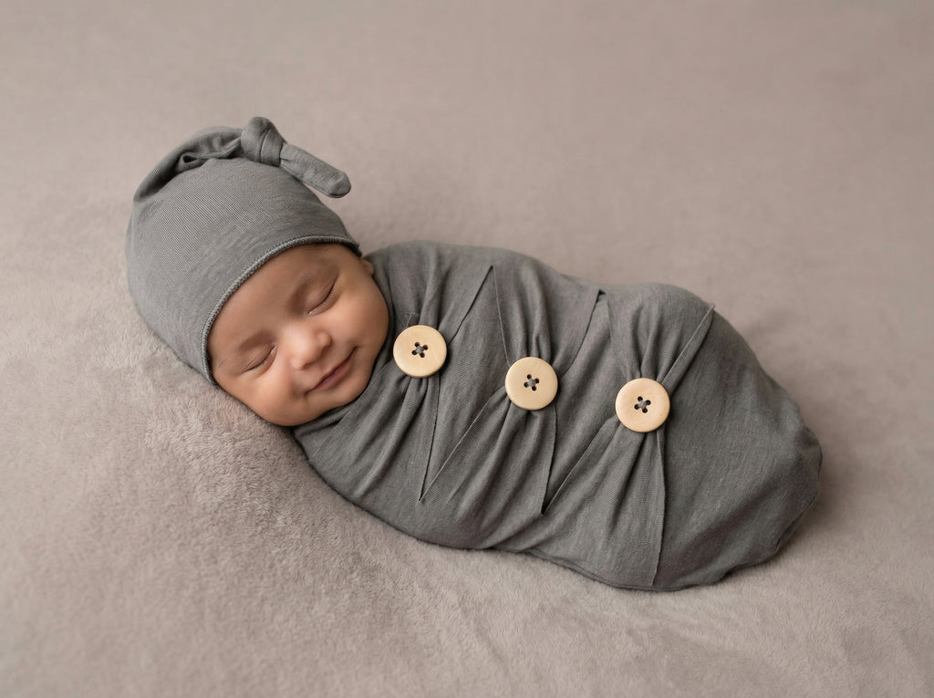 Newborn baby wrapped in a gray swaddle with button details on a beige background