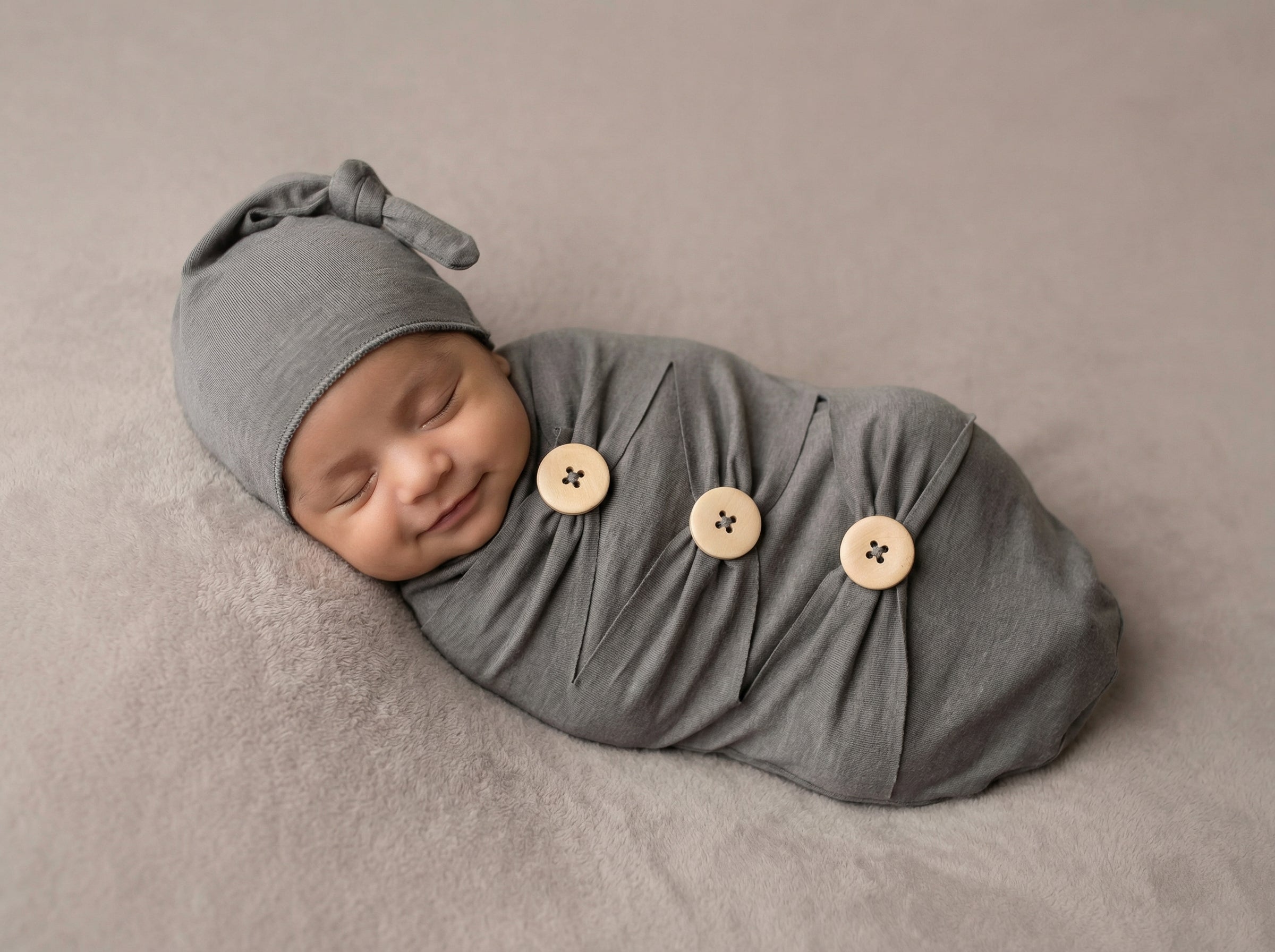 Newborn baby wrapped in a gray swaddle with button details on a beige background