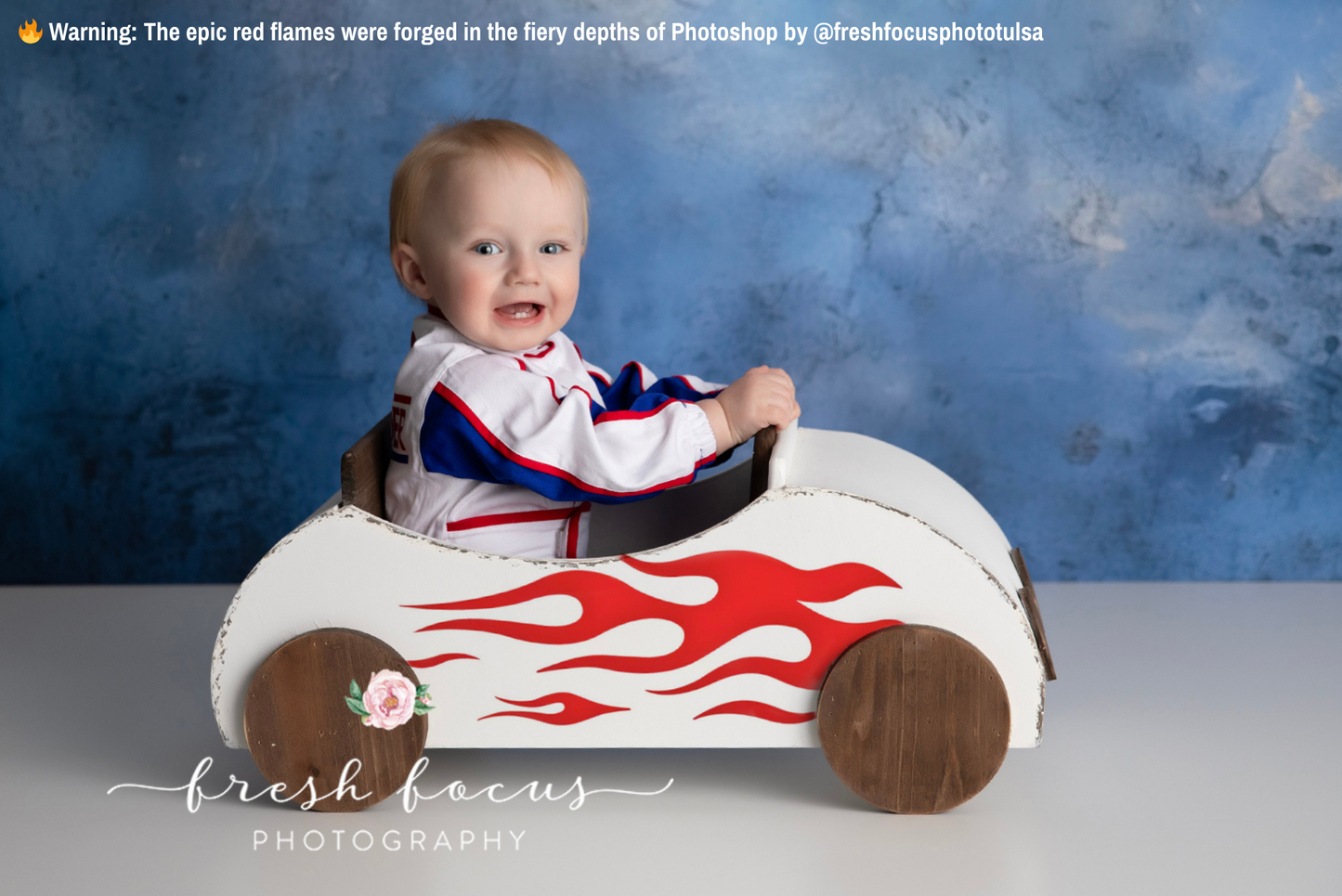 A smiling baby sits in a VW Beetle Cabrio wooden car newborn photography prop, painted white with red flame decals and brown wheels, wearing a race car-themed outfit. 