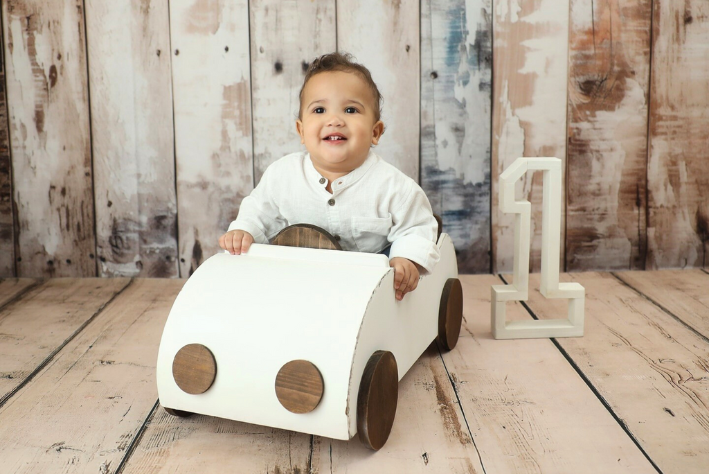 Smiling baby in white shirt sitting in a white wooden car newborn photography prop with rustic backdrop.