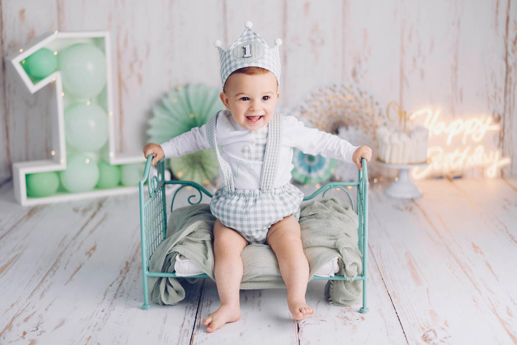 Smiling toddler in a birthday photoshoot sits on a green metallic wire vintage bed, styled as a newborn photography prop. The setup includes soft green fabric, a giant "1" with balloons, and a "Happy Birthday" neon sign.