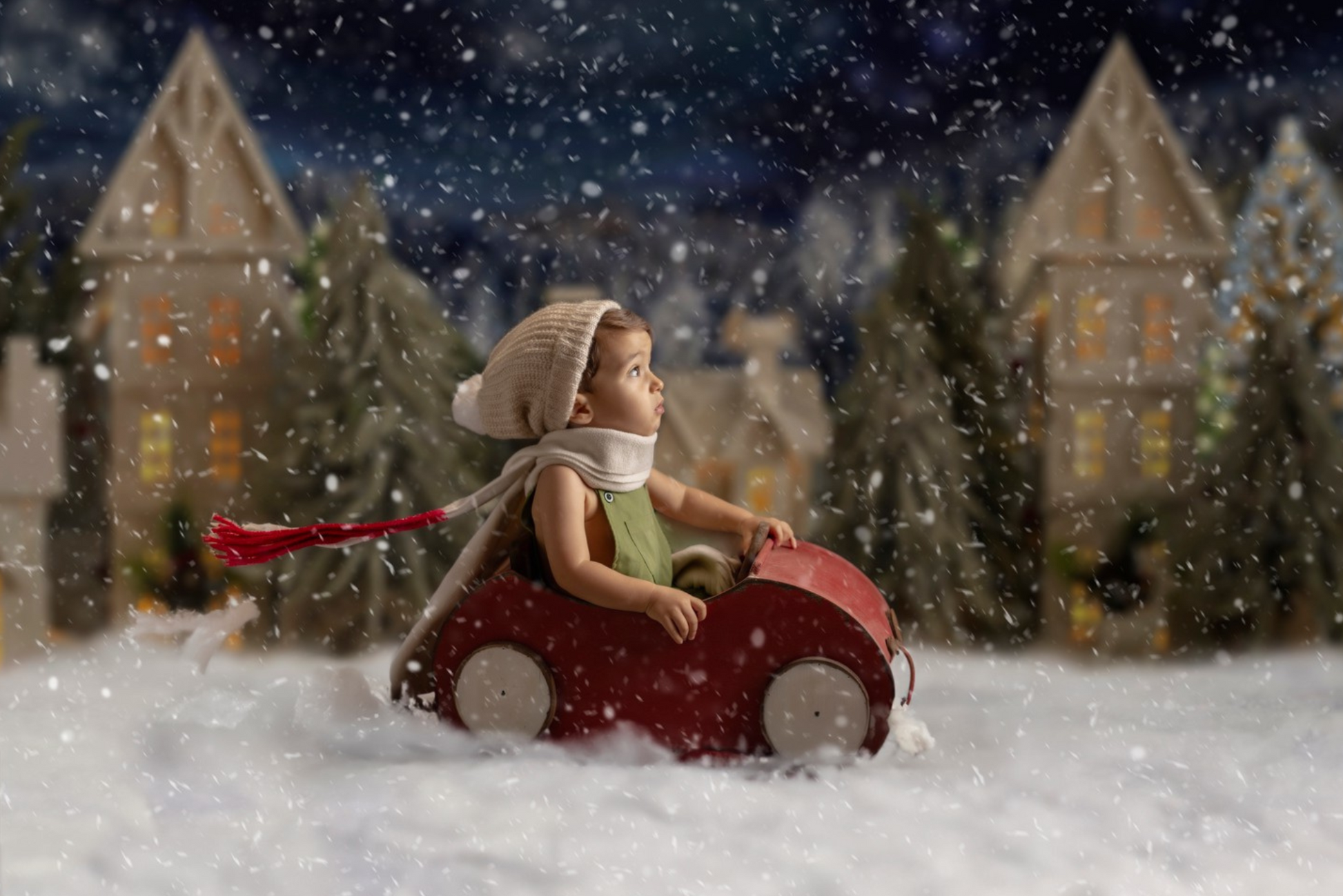 A toddler in a cozy hat and scarf sits in a red wooden toy car, surrounded by falling snow, with a charming village backdrop. Newborn photography prop.