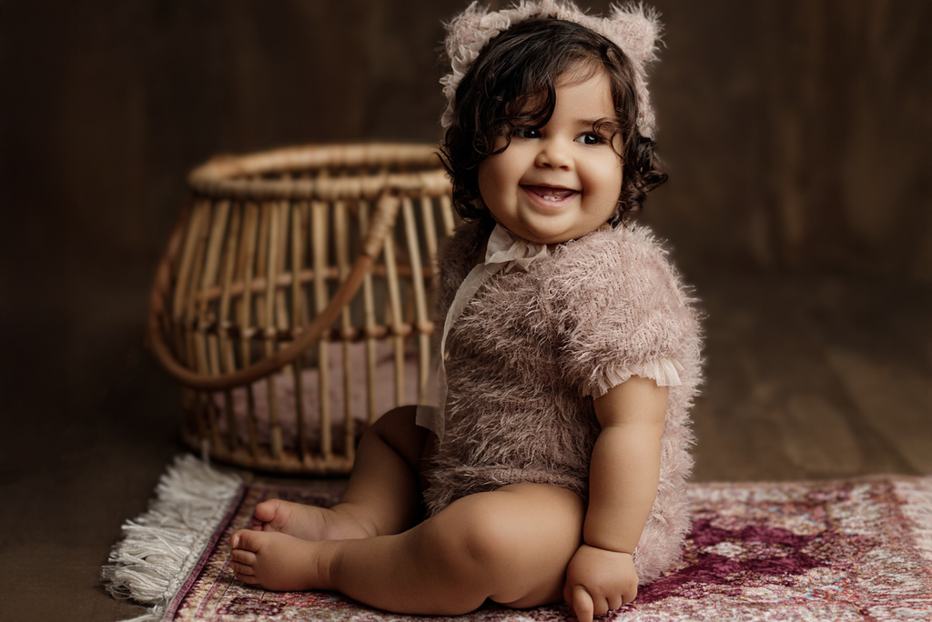 A smiling baby sits on a vintage rug, dressed in a fuzzy bear outfit, with the Woven Wonders rattan basket as a charming newborn photography prop in the background.