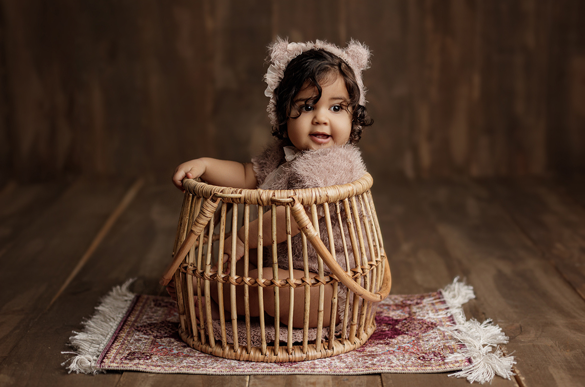 Adorable baby sits in a Woven Wonders rattan basket, wearing a fuzzy bear-themed outfit. The cozy newborn photography prop rests on a vintage rug, creating a warm, rustic setting with soft lighting.