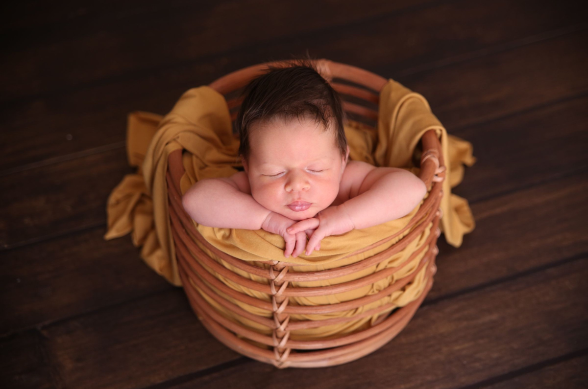 Newborn photography prop featuring a rustic rattan bowl, lined with a mustard-colored wrap. The sleeping baby rests peacefully, head propped on tiny hands, against a warm wooden floor backdrop.