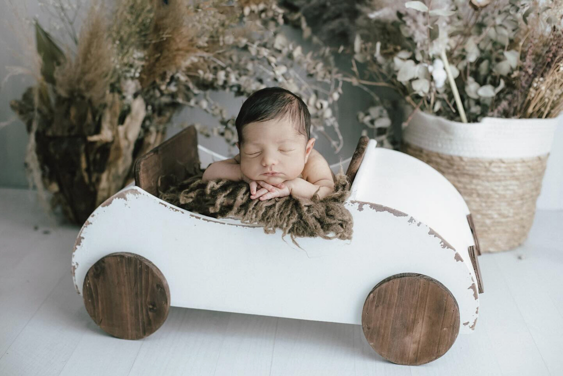 A sleeping newborn rests on a soft brown blanket inside a vintage-style white wooden toy car, with rustic decor in the background. Newborn photography prop.