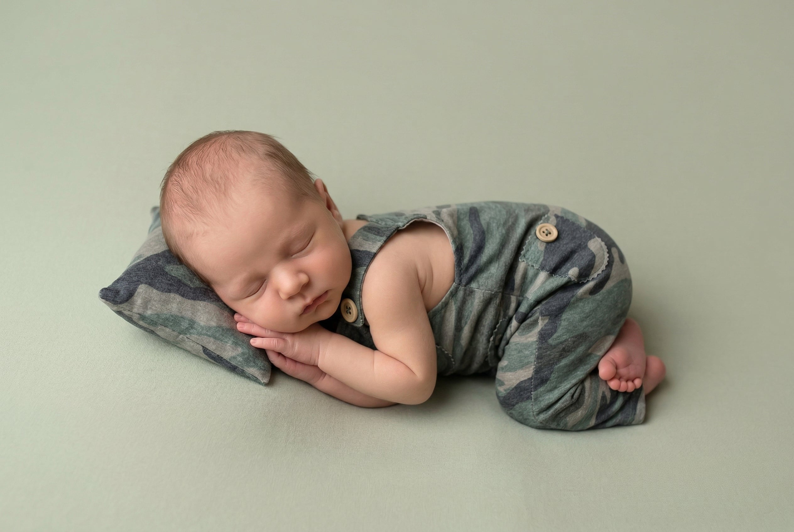 Newborn baby camouflage outfit sleeping on a green blanket with a neutral background