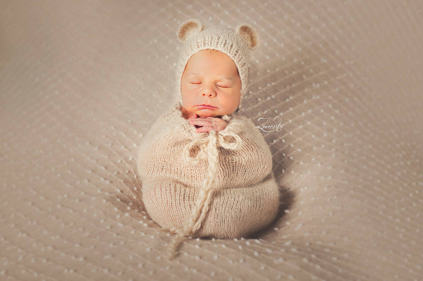 Serene newborn in a beige mohair bear bonnet and sack, resting on a textured cream backdrop, exemplifying a cozy and peaceful newborn photography prop setup.