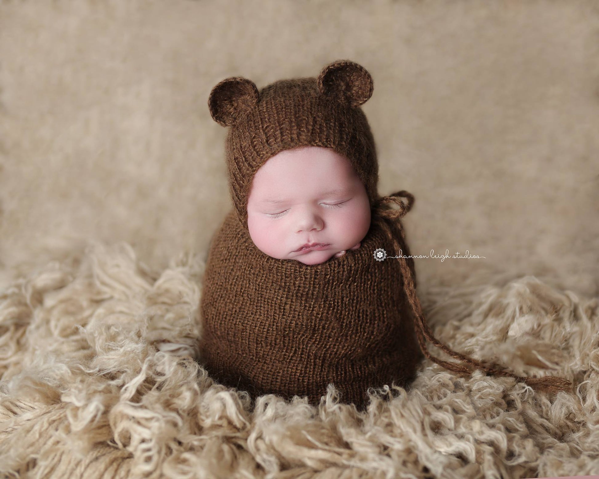 Serene newborn in a beige mohair bear bonnet and sack, resting on a textured cream backdrop, exemplifying a cozy and peaceful newborn photography prop setup.