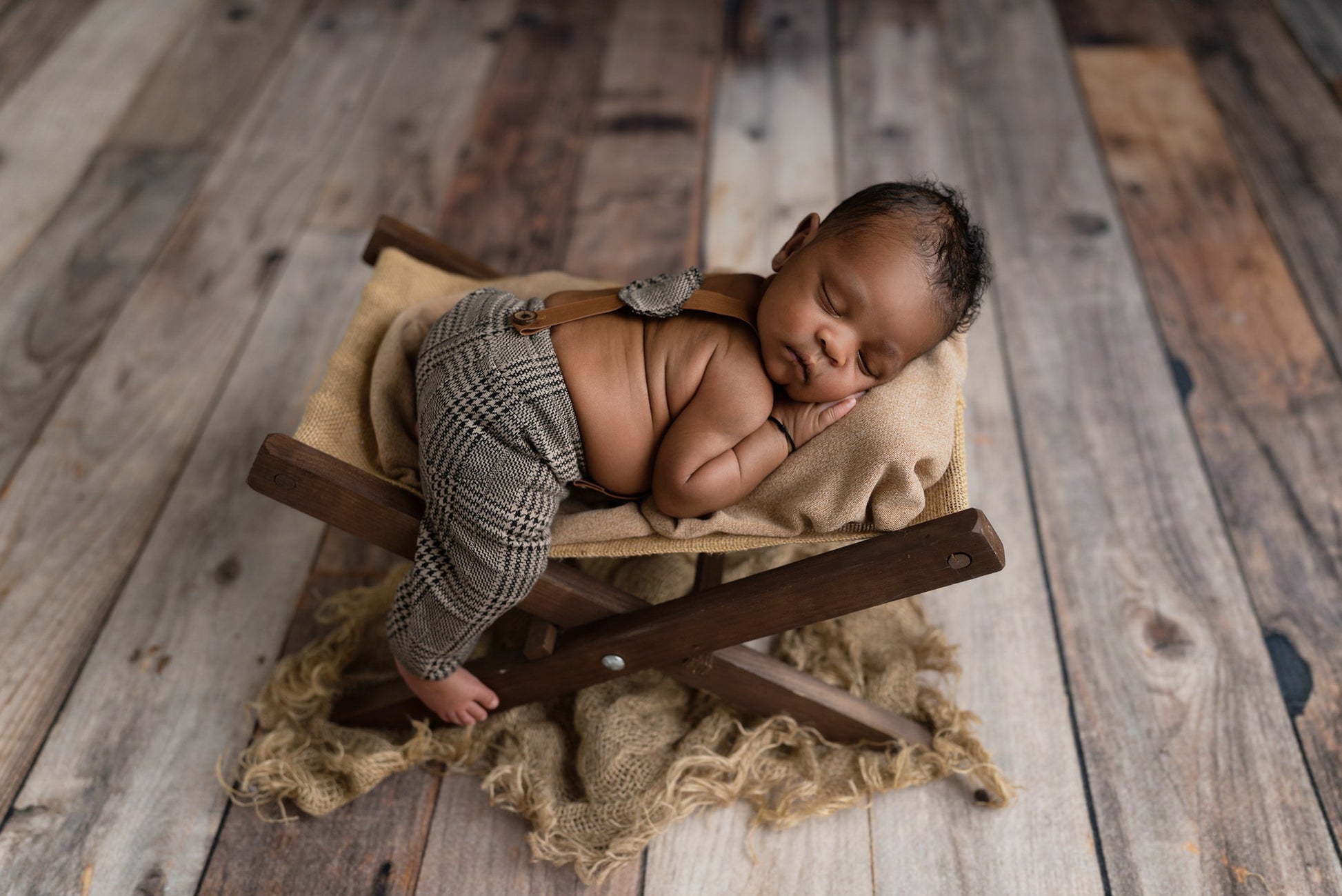 Newborn photography prop: Baby in houndstooth cap and suspenders sleeps on a rustic wooden chair with cozy knit blankets and a soft brown pillow.