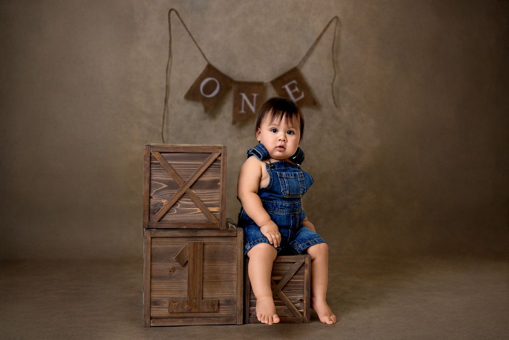 Baby in denim overalls sits on rustic wooden cake smash boxes, a charming milestone photography session prop. One year old.