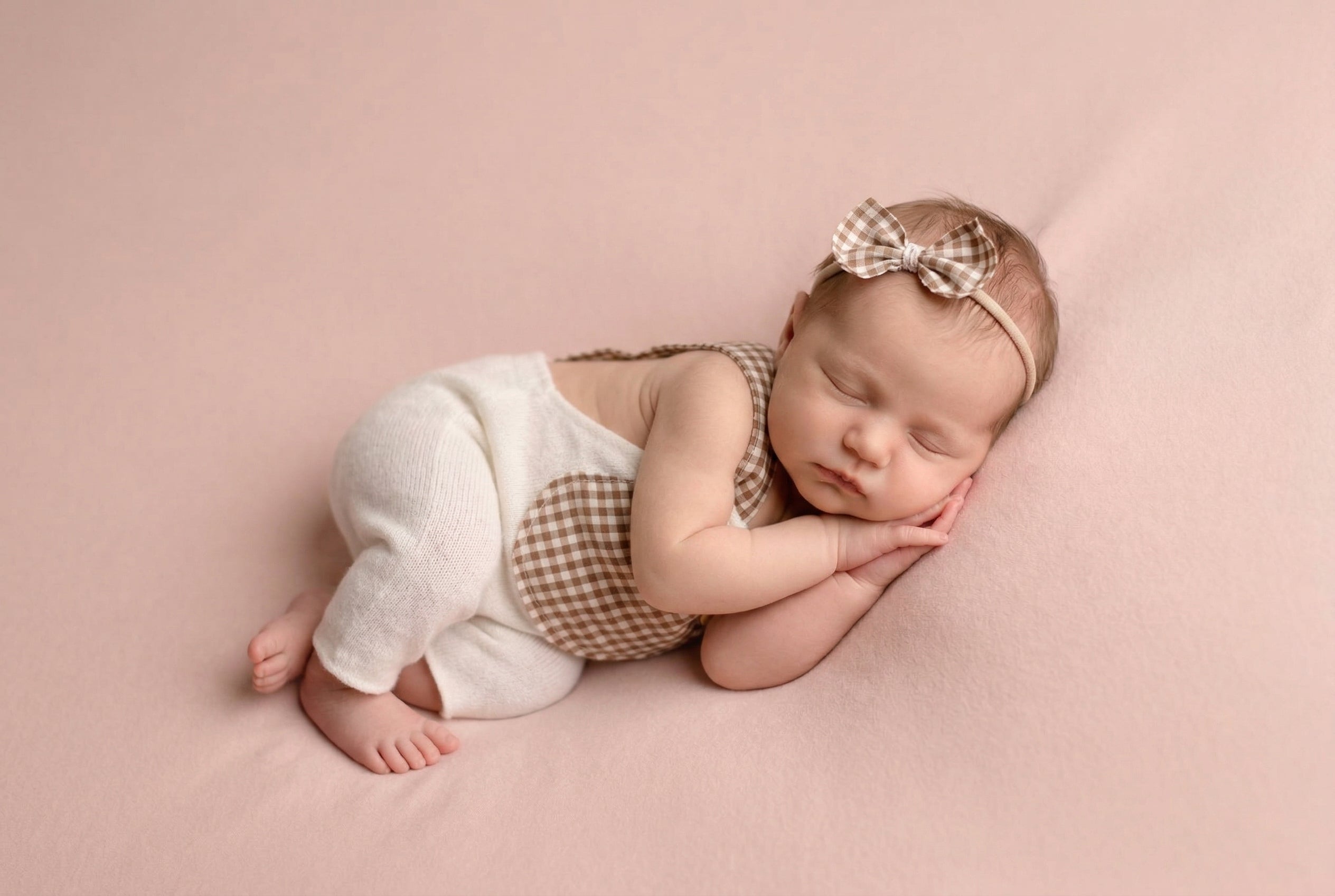Newborn baby sleeping on a pink blanket wearing a checkered outfit and headband.