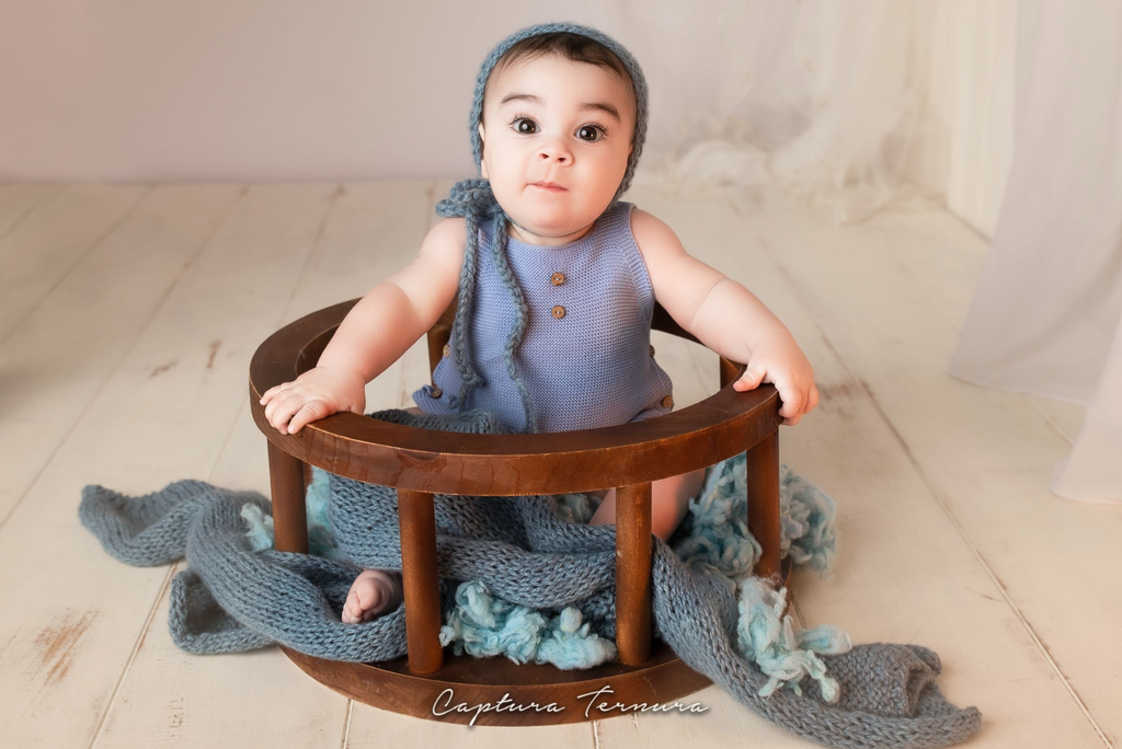 Baby in blue knit outfit and bonnet sitting in a wooden halo seat newborn photography prop with blue wrap.