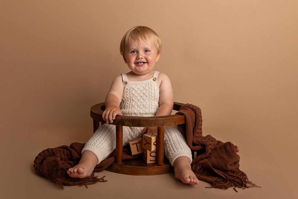 Smiling toddler in cream knit romper sitting in wooden round seat newborn photography prop with brown blanket.