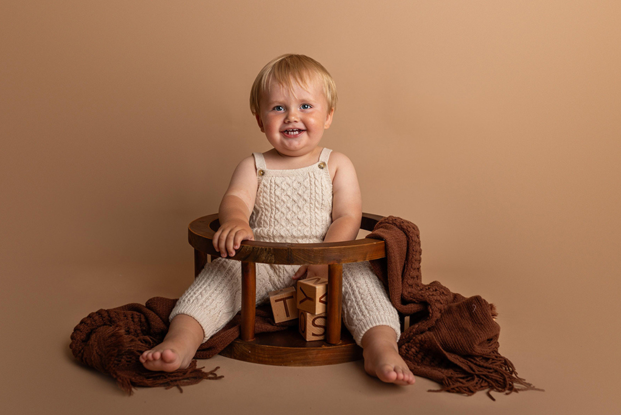 Smiling toddler in cream knit romper sitting in wooden round seat newborn photography prop with brown blanket.