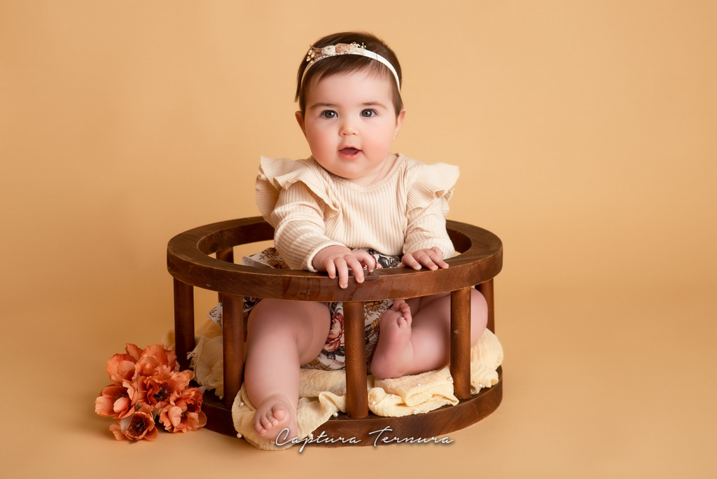Baby girl in ruffled top and floral skirt sitting in wooden halo seat newborn photography prop with peach flowers.