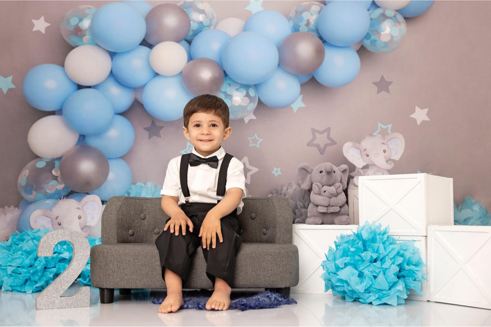 Toddler sitting on a grey couch with a blue balloon arch, plush elephants, and number '2' newborn photography prop.