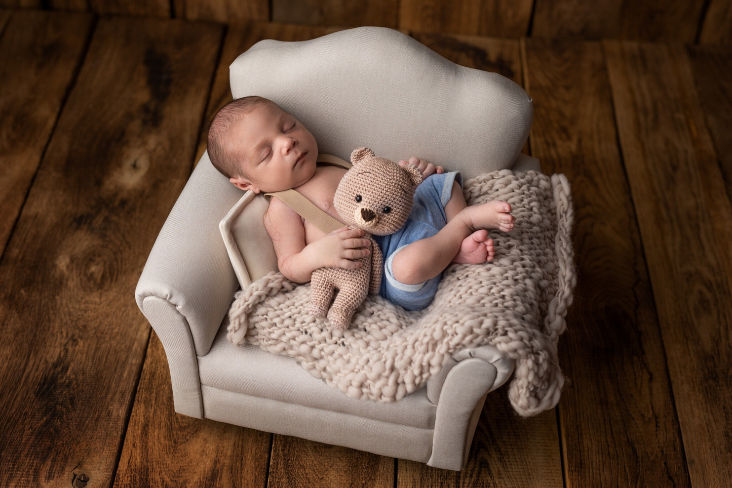 Newborn asleep on a mini sofa with a teddy bear, using a chunky knit blanket as a cozy photography prop.