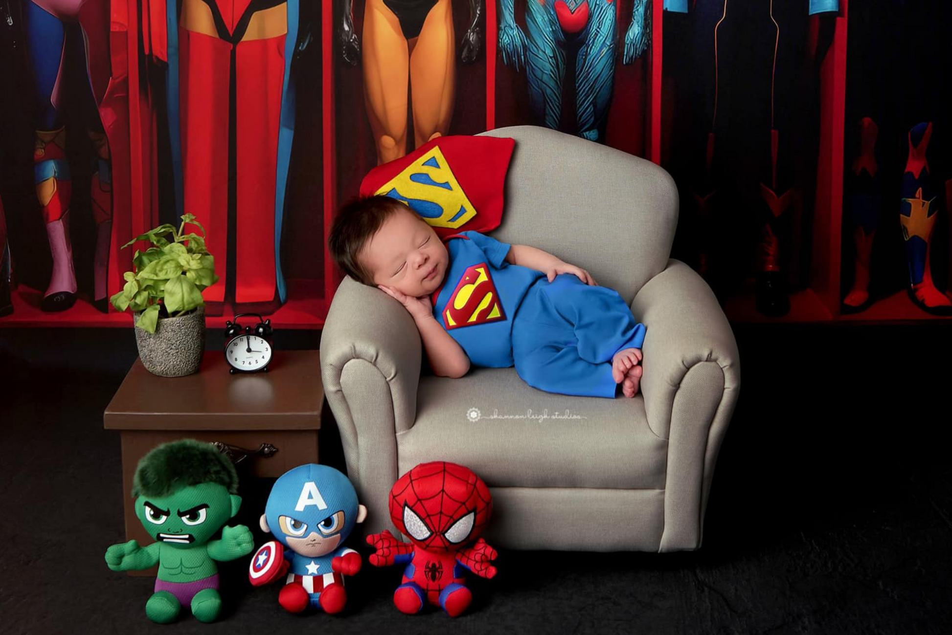 Newborn photography prop setup featuring a gray mini couch with a baby in a Superman outfit, surrounded by superhero plush toys.