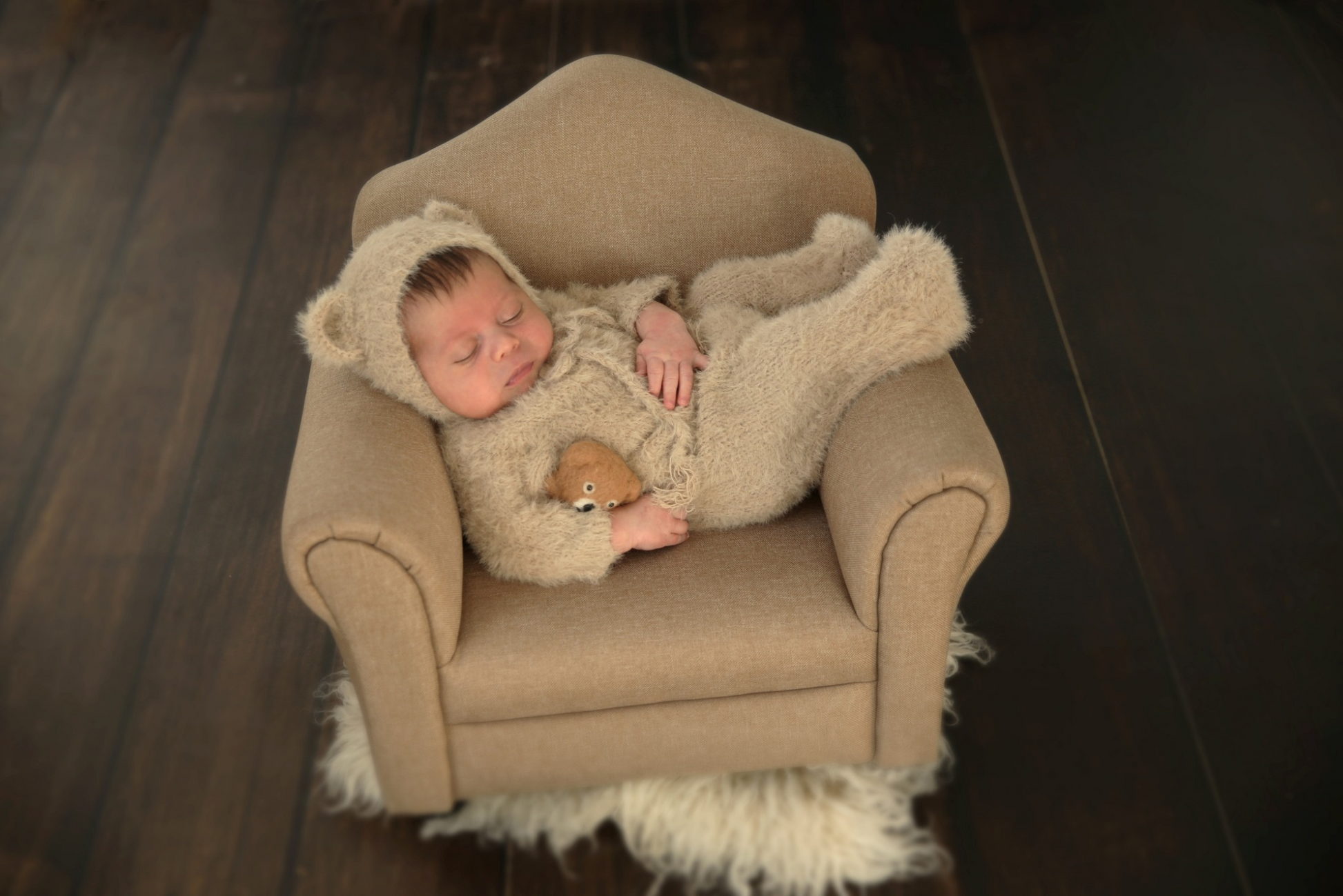 Sleeping baby on a light brown mini sofa, wearing a bear romper and bonnet as a newborn photography prop.