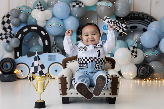 Adorable race car-themed newborn photography setup featuring a plush mini sofa as a cozy newborn photography prop. The baby, dressed in a racing outfit, sits excitedly on the cream and brown upholstered chair, surrounded by a checkered flag, a golden trophy, and a blue balloon backdrop with race car details. The perfect prop for a themed milestone session!