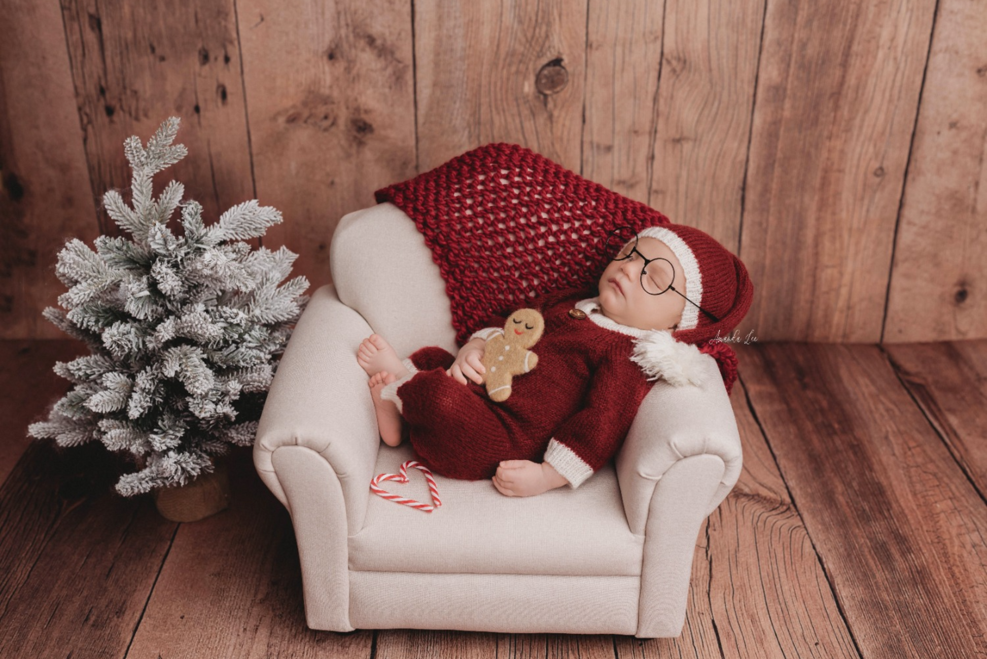 Newborn photography prop featuring a tiny cream sofa couch, with a baby dressed in a red Santa outfit and glasses. The baby holds a gingerbread toy while resting near a frosted mini Christmas tree.