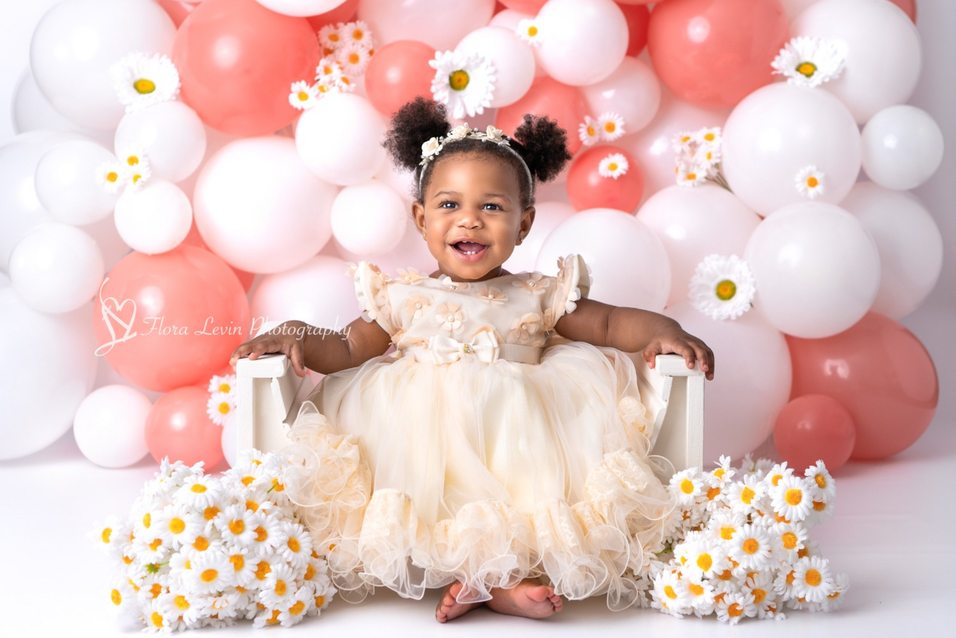 miling baby girl in a delicate cream dress, sitting on a white Arch Lounger for newborn photography, surrounded by white and coral balloons and daisies. The Arch Lounger is designed for comfortable posing in baby photo sessions, providing both support and aesthetic appeal in a soft, minimalist style.