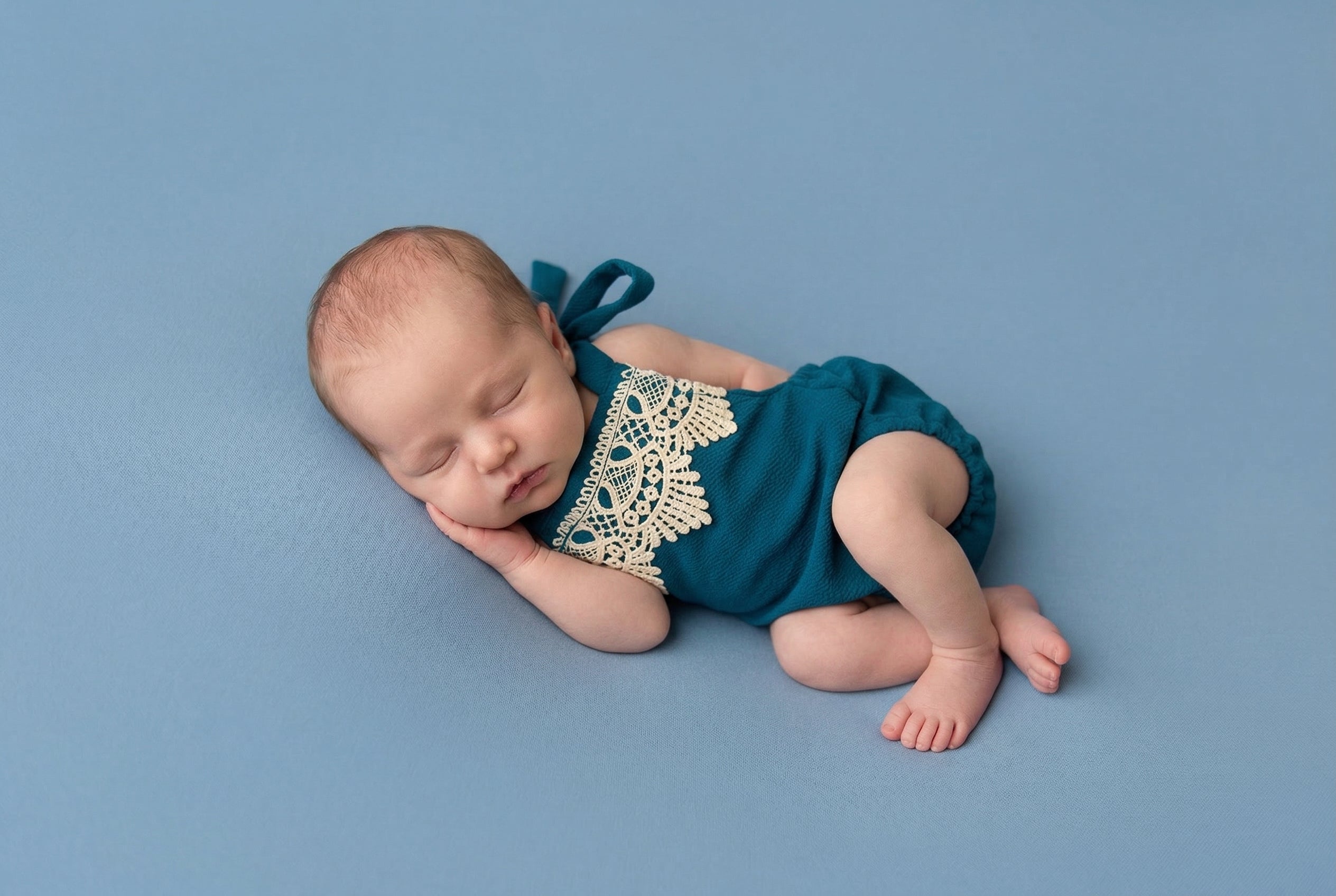 Newborn baby in a teal outfit with lace details on a blue background