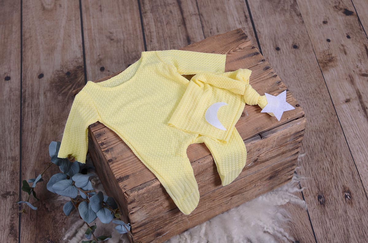 Yellow waffle fabric footed pajamas and matching hat with moon and stars, displayed as a newborn photography prop on a rustic wooden surface.