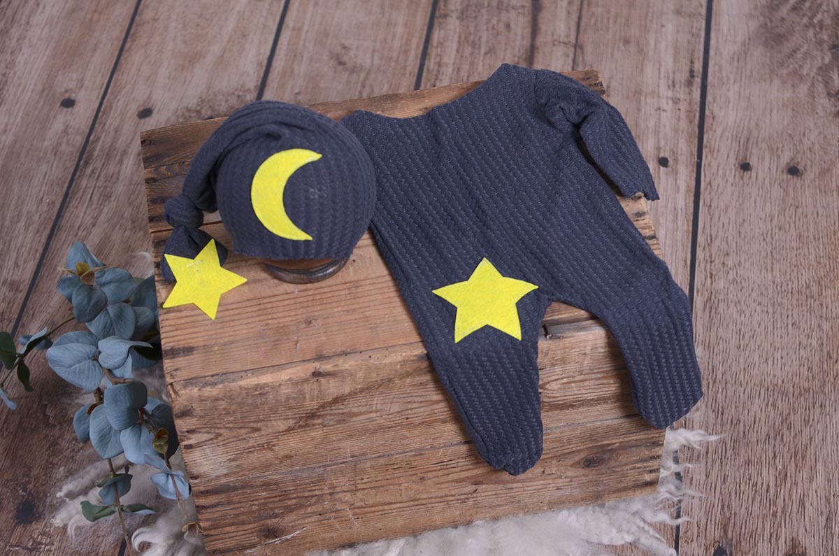Blue waffle fabric footed pajamas and matching hat with moon and stars, displayed as a newborn photography prop on a rustic wooden surface.