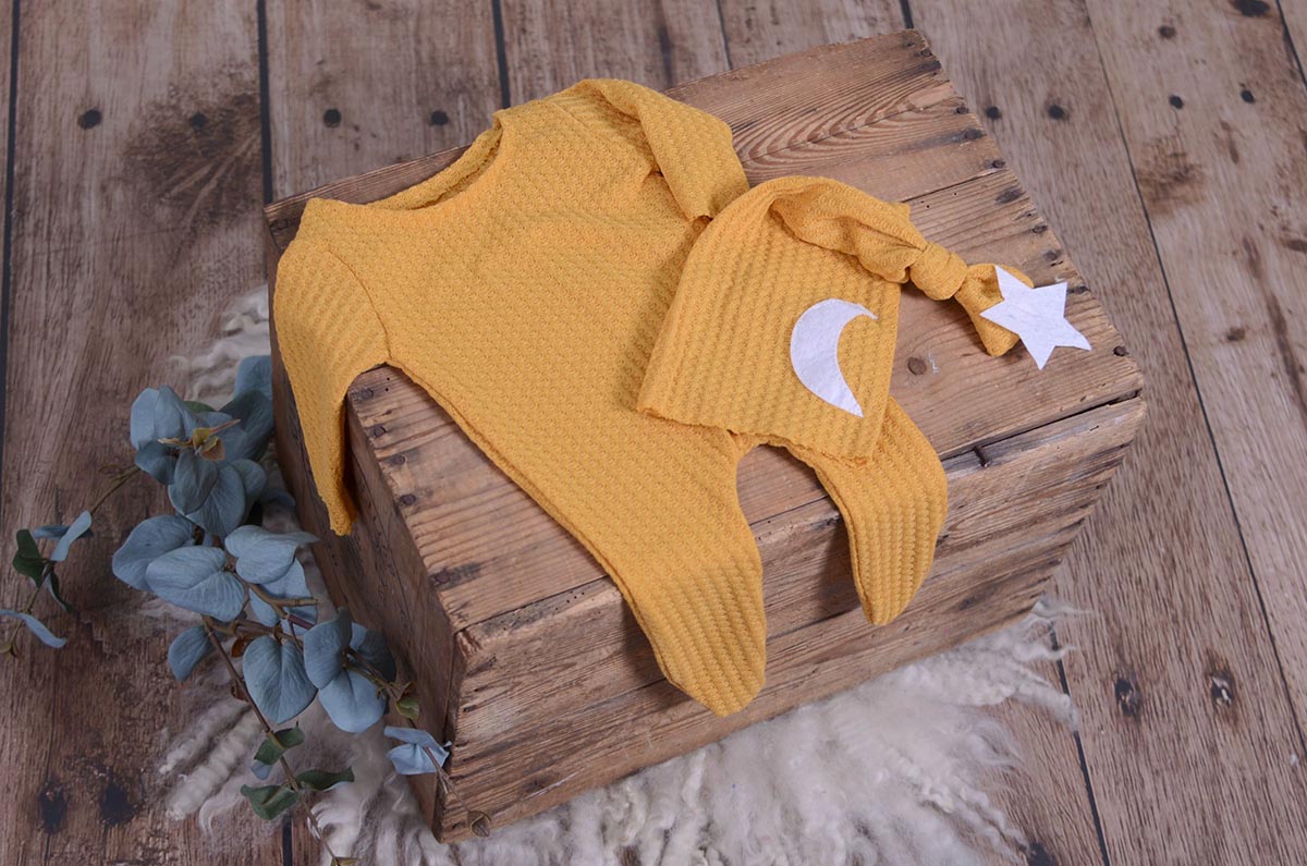 Mustard waffle fabric footed pajamas and matching hat with moon and stars, displayed as a newborn photography prop on a rustic wooden surface.