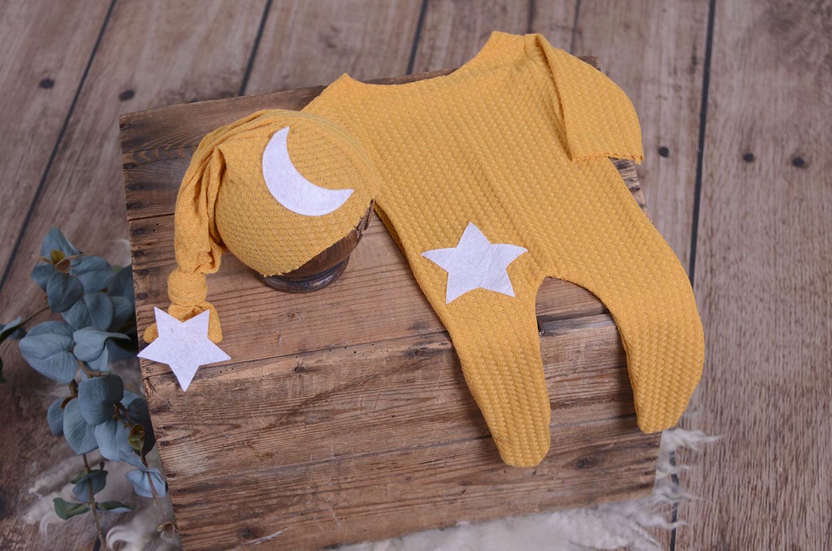 Mustard waffle fabric footed pajamas and matching hat with moon and stars, displayed as a newborn photography prop on a rustic wooden surface.