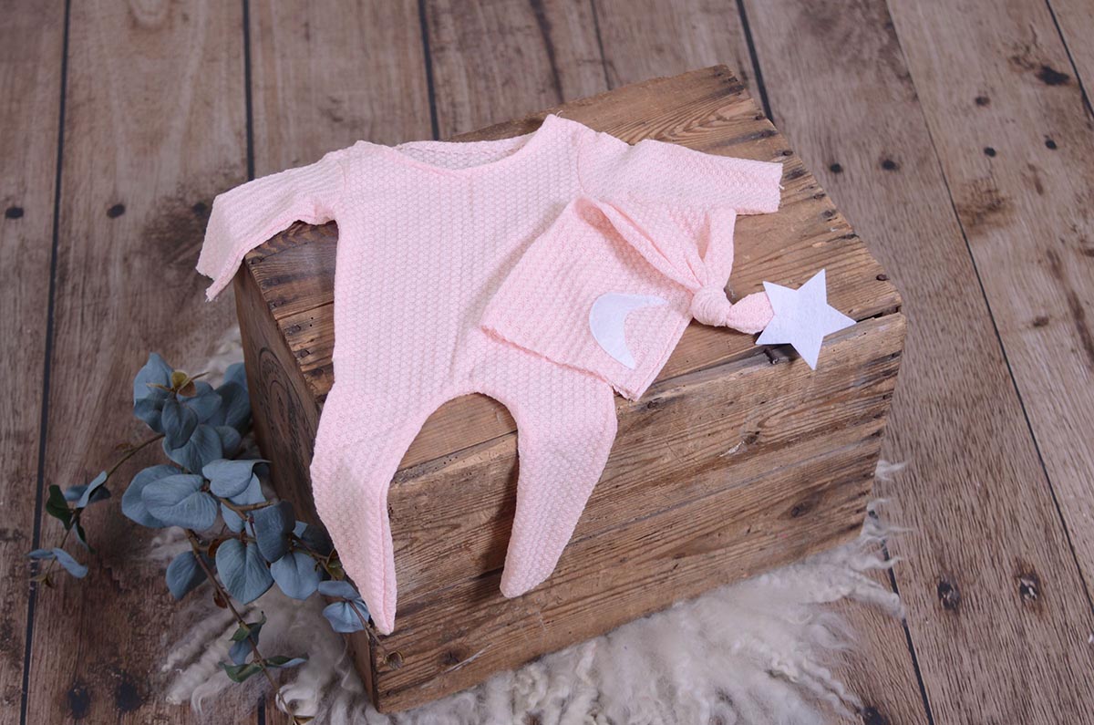 Pink waffle fabric footed pajamas and matching hat with moon and stars, displayed as a newborn photography prop on a rustic wooden surface.