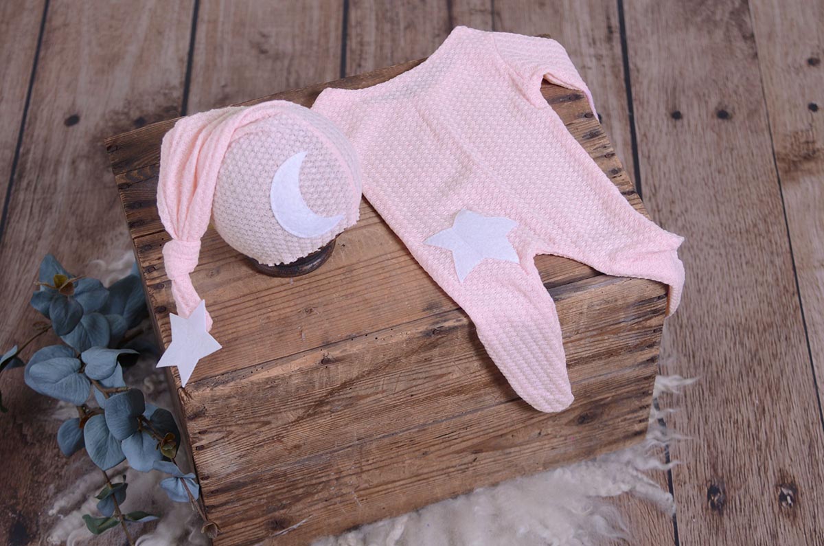 Pink waffle fabric footed pajamas and matching hat with moon and stars, displayed as a newborn photography prop on a rustic wooden surface.