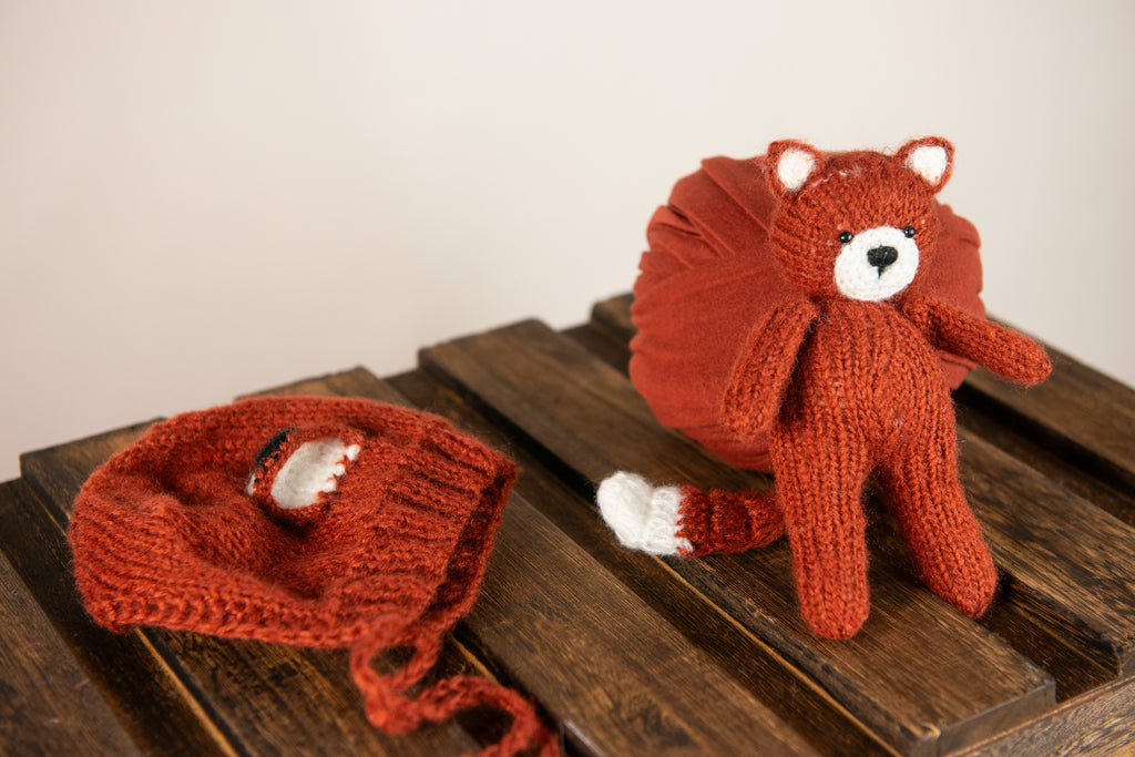 Newborn snuggled in dark orange wrap, with fox bonnet and matching plush, ideal for a warm-toned newborn photography prop setup.