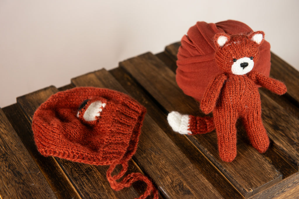 Newborn snuggled in dark orange wrap, with fox bonnet and matching plush, ideal for a warm-toned newborn photography prop setup.