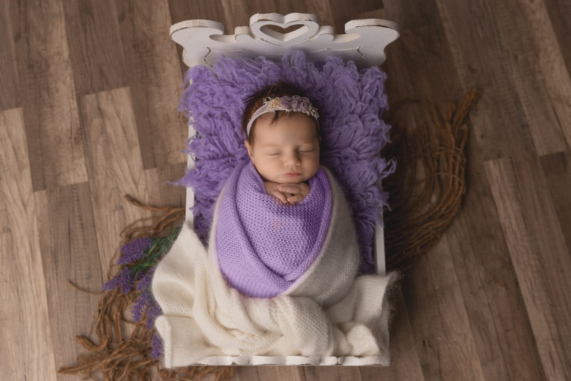 Newborn peacefully sleeping in a white vintage bed, swaddled in a lavender knit wrap atop a fluffy purple layer. Newborn photography prop serene scene. The headboard of the vintage bed features a delicate, heart-shaped cutout at the center.