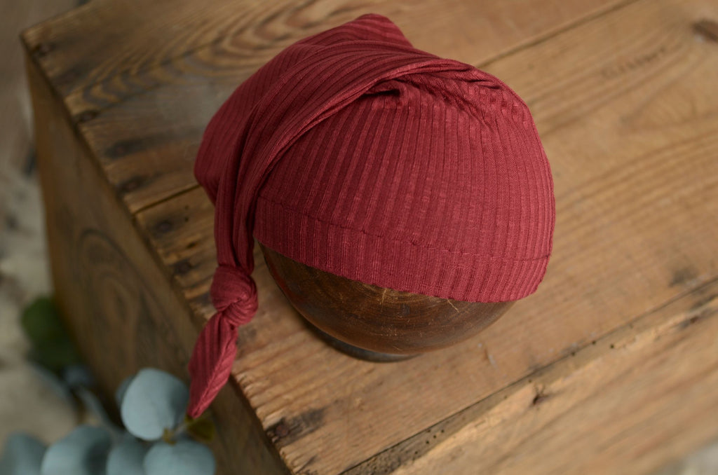 Ribbed knot sleepy cap newborn photography prop in deep burgundy, styled on a rustic wooden bowl.